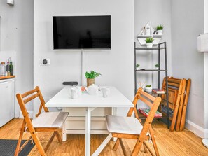 A modern kitchen with a white table, two wooden chairs, a TV mounted on the wall, and a small shelf with plants. - No.3 West Beach Apartments - UK52003 (Portrush)