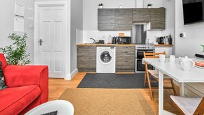 A compact living space with a kitchenette, a washing machine, a dining area, and a red sofa. - No.3 West Beach Apartments - UK52003 (Portrush)