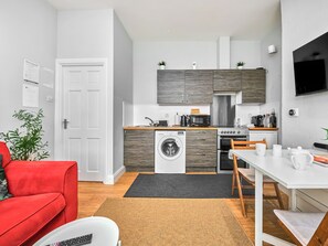 A compact living space with a kitchenette, a washing machine, a dining area, and a red sofa. - No.3 West Beach Apartments - UK52003 (Portrush)