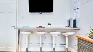 Ett modernt kök med en platt-TV monterad ovanför en bänkskiva, tre vita barstolar, en kaffemaskin och en liten krukväxt.