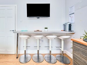 Eine moderne Küche mit Flachbildfernseher über einer Arbeitsplatte, drei weiße Barhocker, einer Kaffeemaschine und einem kleinen Topf mit Pflanze.