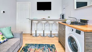 Ett kompakt vardagsrum med tvättmaskin, en liten bardisk, en soffa och en färgglad matta.