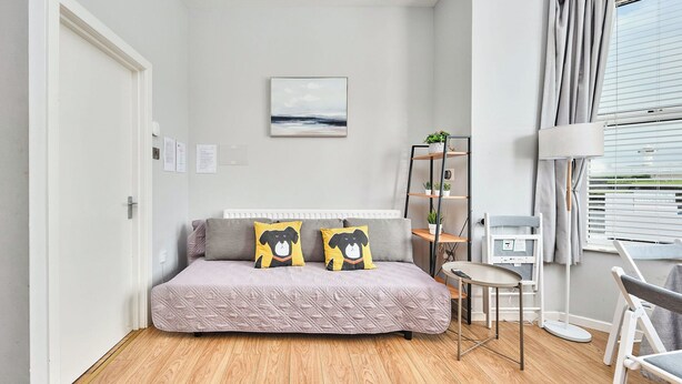 A room with a bed, a small table, a radiator, a shelf with plants, and a window with curtains. - No.1 Dunluce Park Apartments - UK52007 (Portrush)
