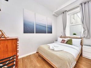 Ein Schlafzimmer mit einem Bett, einem Nachttisch, Vorhängen an einem Fenster und einem Wandbild.