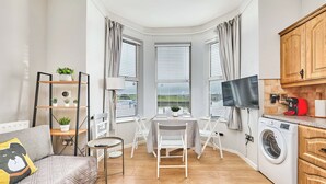 A compact living space with a sofa, a small dining table, a TV, a washing machine, and a kitchen area with cabinets. - No.1 Dunluce Park Apartments - UK52007 (Portrush)