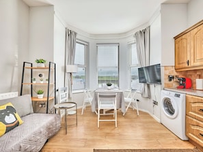 Ein kompakter Wohnraum mit Sofa, einem kleinen Esstisch, Fernseher, Waschmaschine und Küchenzeile mit Schränken.
