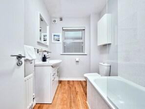 Ein modernes Badezimmer mit einem weißen Waschbecken, Spiegel und WC, Holzboden und einer Badewanne.