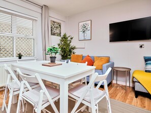 Ein modernes Wohnzimmer mit einem weißen Esstisch, weißen Stühlen, einem blauen Sofa mit orangefarbenen Kissen, einem kleinen runden Tisch und einem an der Wand befestigten Flachbildfernseher.