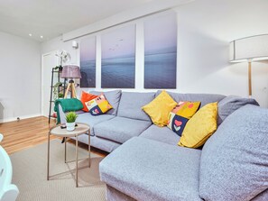 Ein modernes Wohnzimmer mit einem grauen Sofa, bunten Kissen, einem kleinen Beistelltisch, einer Stehlampe und einem großen Fenster mit Meerblick.