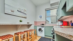 Een kleine keuken met een wasmachine, houten krukken, een aanrecht en een raam met zonwering.