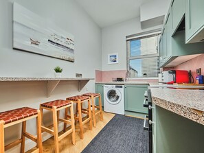 A compact kitchen with a washing machine, wooden stools, a countertop, and a window with blinds. - No.3 Dunluce Apartments - UK52009 (Portrush)