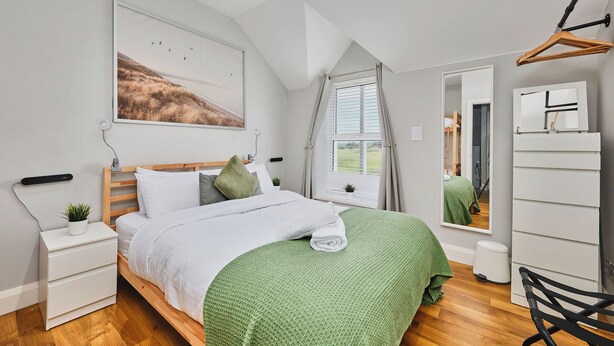 Ein modernes Schlafzimmer mit Bett, Nachttischen, einer grünen Decke, einem Fenster mit Vorhängen, einem Spiegel und einer Kleiderstange.