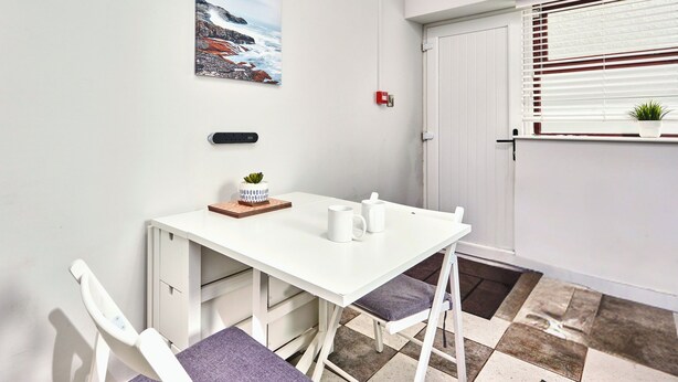 Een klein, wit eetgedeelte met een tafel en stoelen, een schilderij aan de muur, een stopcontact, een rood noodlicht en een raam met jaloezieën.