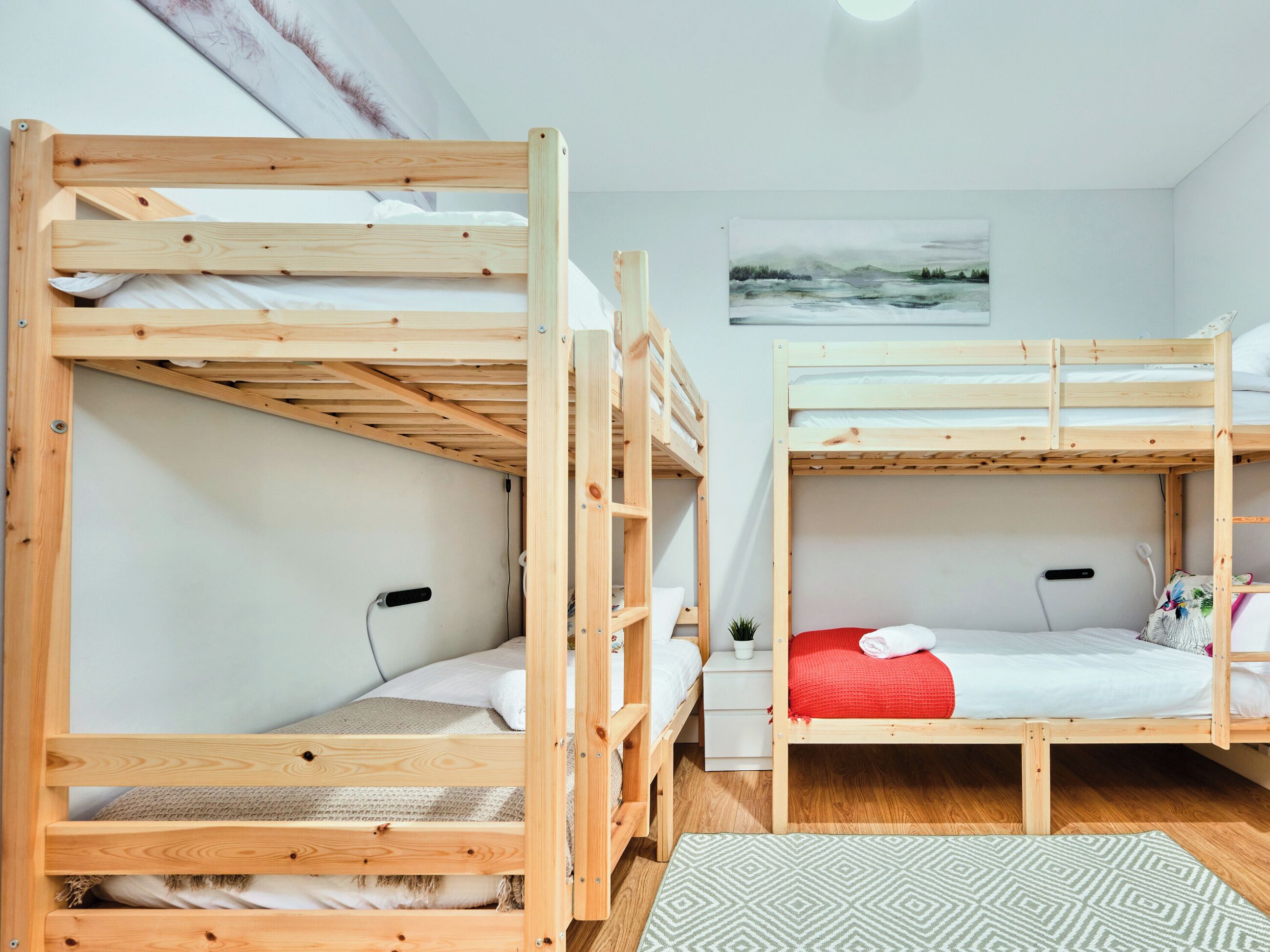 Ein Zimmer mit zwei Etagenbetten, einem kleinen Nachttisch, einer wandmontierten Steckdose und einem gerahmten Bild an der Wand.