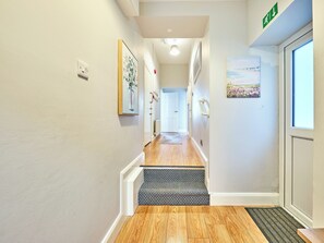 A hallway with a wooden floor, a black and white patterned mat at the entrance, and a door with a green exit sign. - No.2 Dunluce Green Apartments - UK52015 (Portrush)
