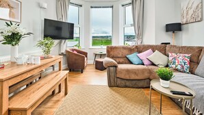 Moderni olohuone, jossa on suuri ikkuna, litteänäyttöinen televisio, ruskea sohva värikkäillä koristetyynyillä, puinen sohvapöytä ja penkki.