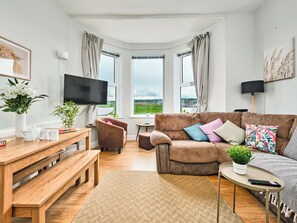 A modern living room with a large window, a flat-screen TV, a brown sofa with colorful cushions, a wooden coffee table, and a bench. - No.2 Dunluce Green Apartments - UK52015 (Portrush)
