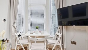A room with a white table, two white chairs, a TV, and a window with blinds. - No.2 Princess Red Apartments - UK52041 (Portrush)