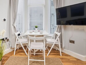 Ein Zimmer mit einem weißen Tisch, zwei weißen Stühlen, einem Fernseher und einem Fenster mit Jalousien.