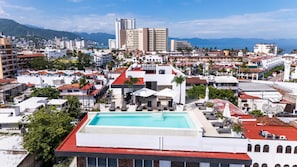 Ein Pool auf dem Dach mit Liegestühlen und Sonnenschirmen, umgeben von Gebäuden und einer Bergkulisse.