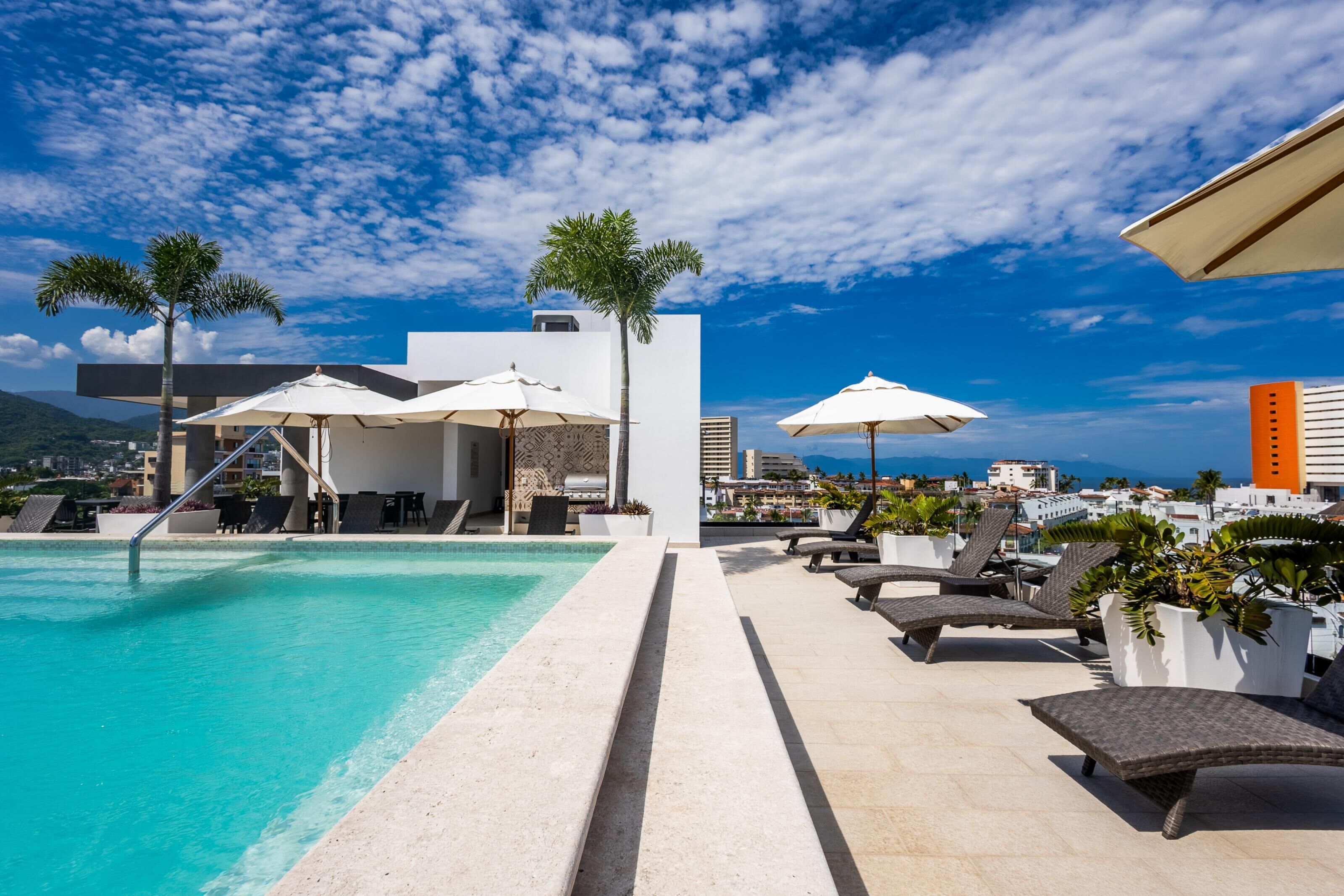 Un espace piscine aménagé sur le toit, avec des chaises longues, des parasols et une vue sur le paysage urbain.