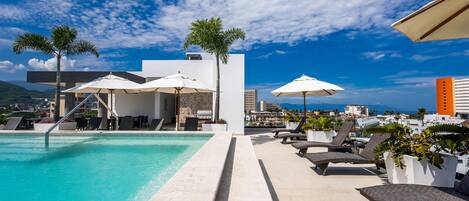 A rooftop pool area with lounge chairs, umbrellas, and a view of the cityscape.