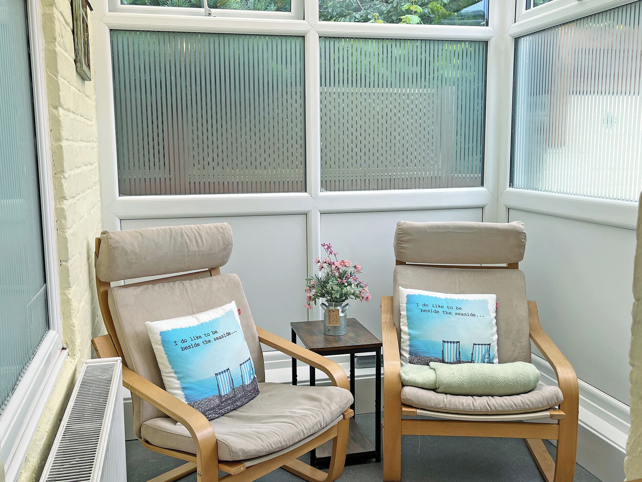 Two wooden chairs with beige cushions and matching pillows, positioned in front of a window with a view of greenery.