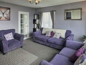 Ein Wohnzimmer mit einem violetten Sofa, einem passenden Sessel und einem Glastisch. Eine Tür mit Milchglasfüllung, ein Bücherregal und ein Spiegel an der Wand sind ebenfalls vorhanden.