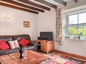 Ein Wohnzimmer mit einer Ledersofa, einem Couchtisch aus Holz mit einer Flasche und einem Glas, einem Fernseher auf einem Holzständer und einem Fenster mit Blick ins Grüne.