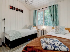 Ein Schlafzimmer mit zwei Betten, einem Fenster mit Vorhängen, einem Nachttisch mit Lampe und einem an der Wand hängenden Kunstwerk.
