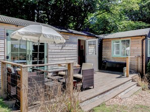 Eine Holzterrasse mit Gartenmöbeln, einem überdachten Sitzbereich und einem kleinen Gebäude mit Tür und Fenstern.