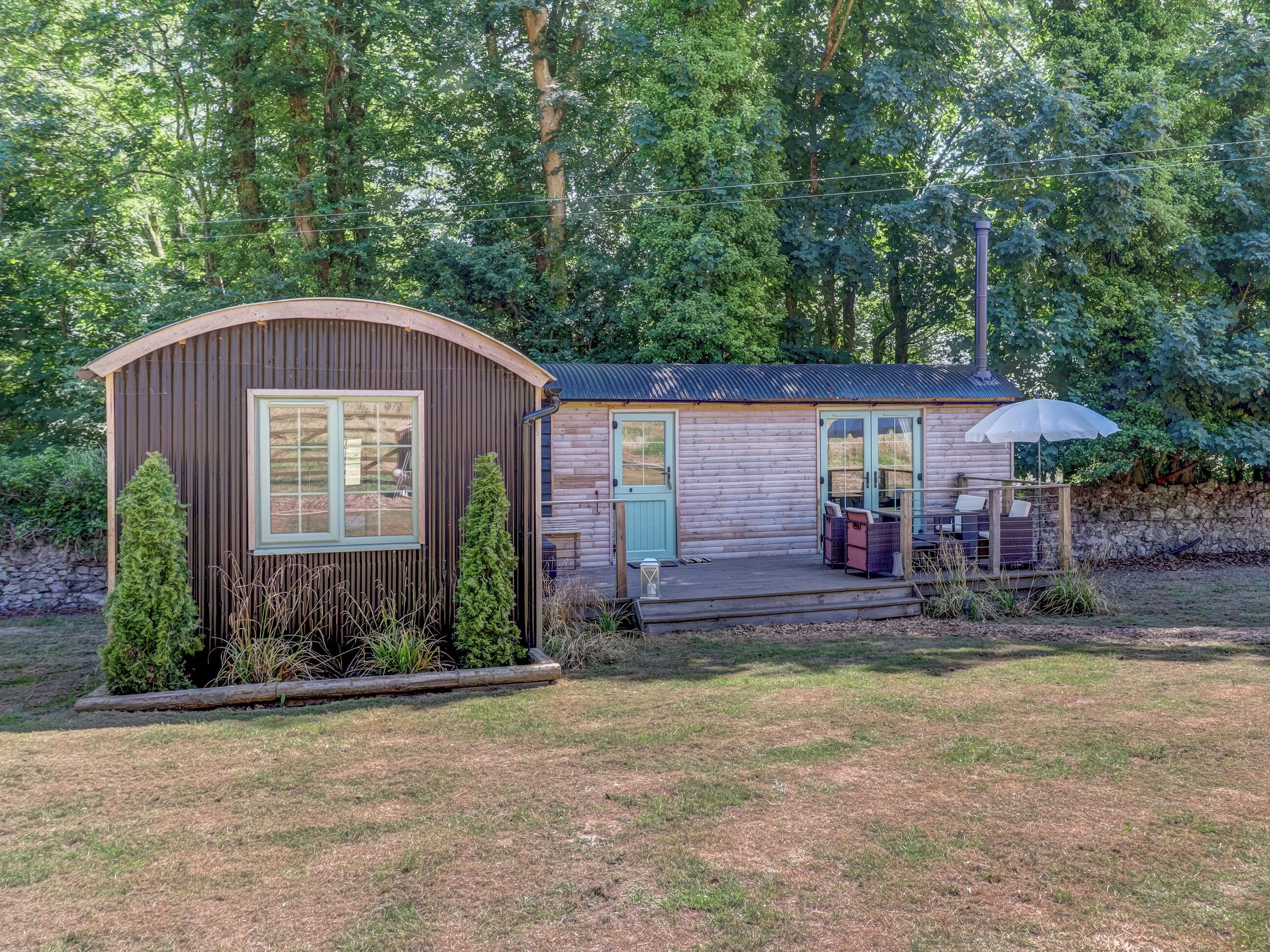 A wooden cabin with a curved roof, a porch with outdoor seating, and a stone wall in the background.