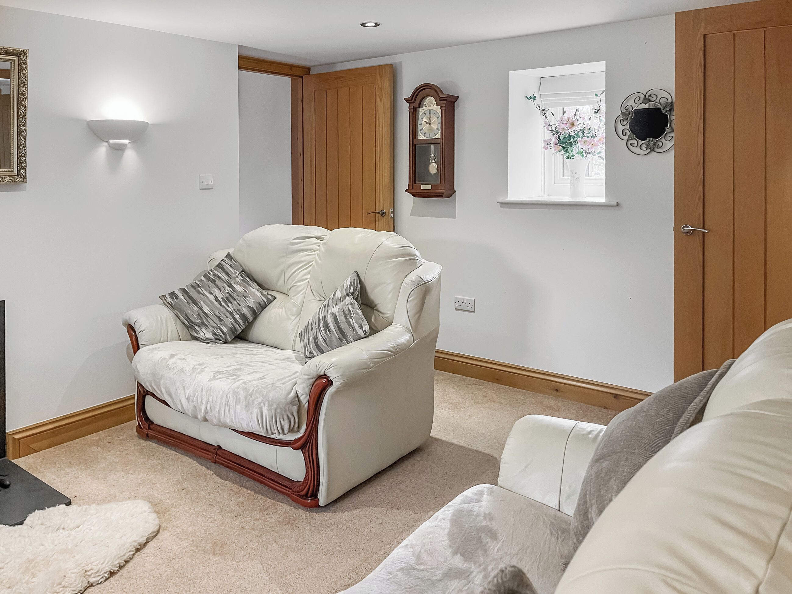 A living room with a cream-colored sofa, a wooden-framed armchair, a fluffy white rug, a wall-mounted clock, and a window with a decorative frame.