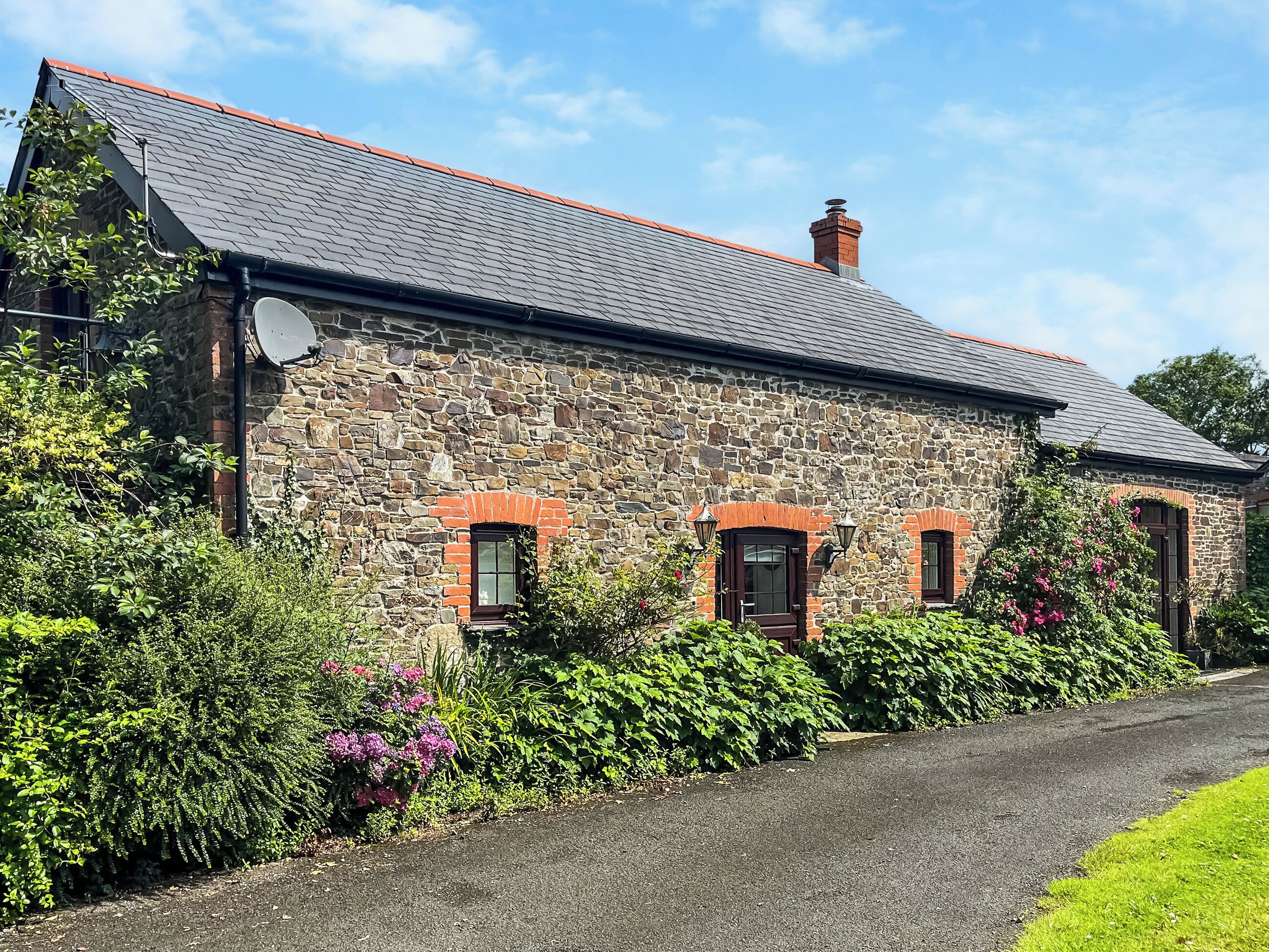 A stone house with a sloped roof and a chimney, surrounded by lush greenery and a paved driveway.