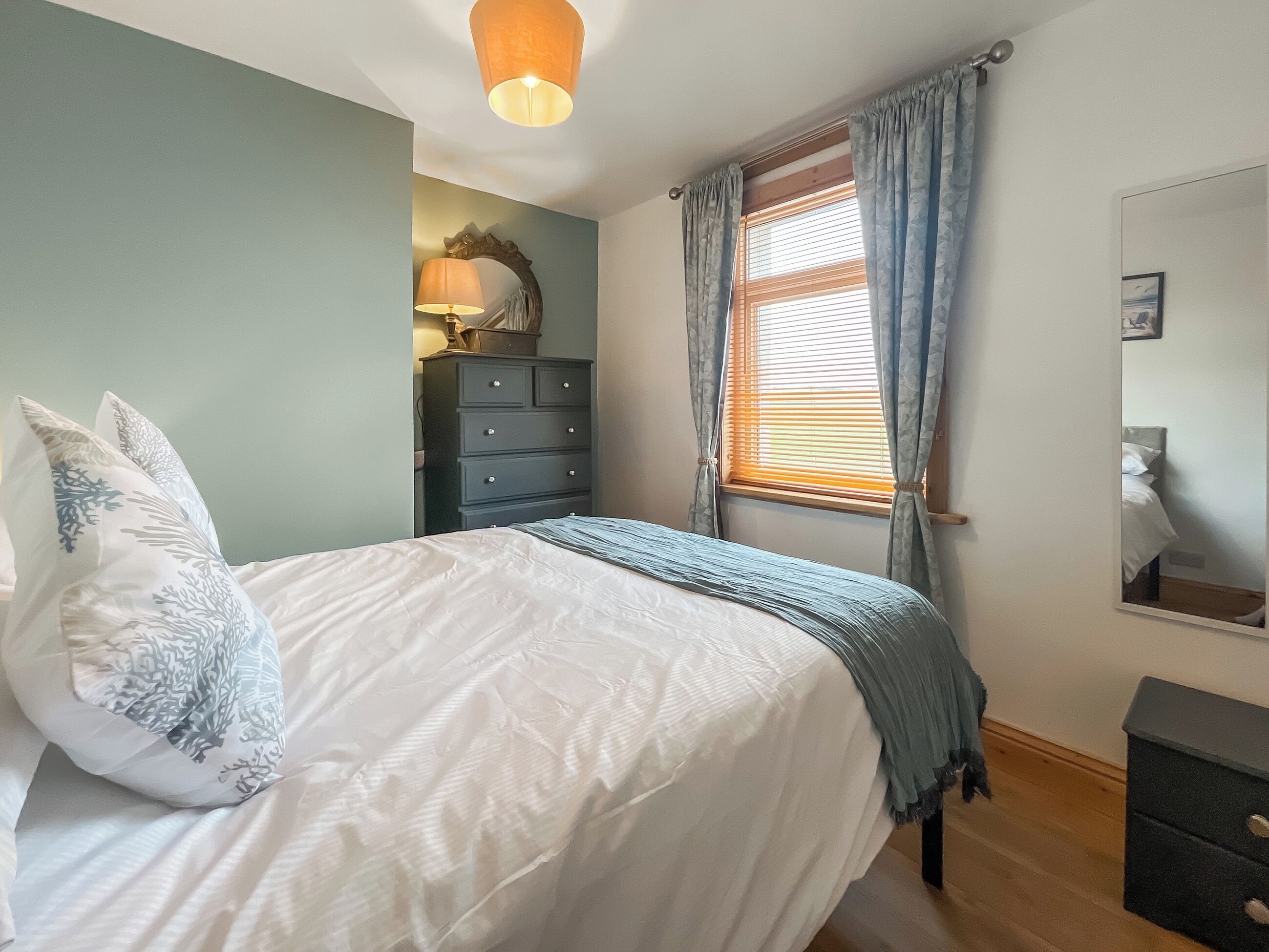 Une chambre à coucher avec un lit, une commode, une lampe et une fenêtre avec des rideaux.