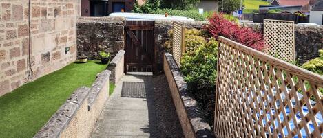 A narrow pathway leading to a gated entrance, flanked by stone walls and a wooden fence.