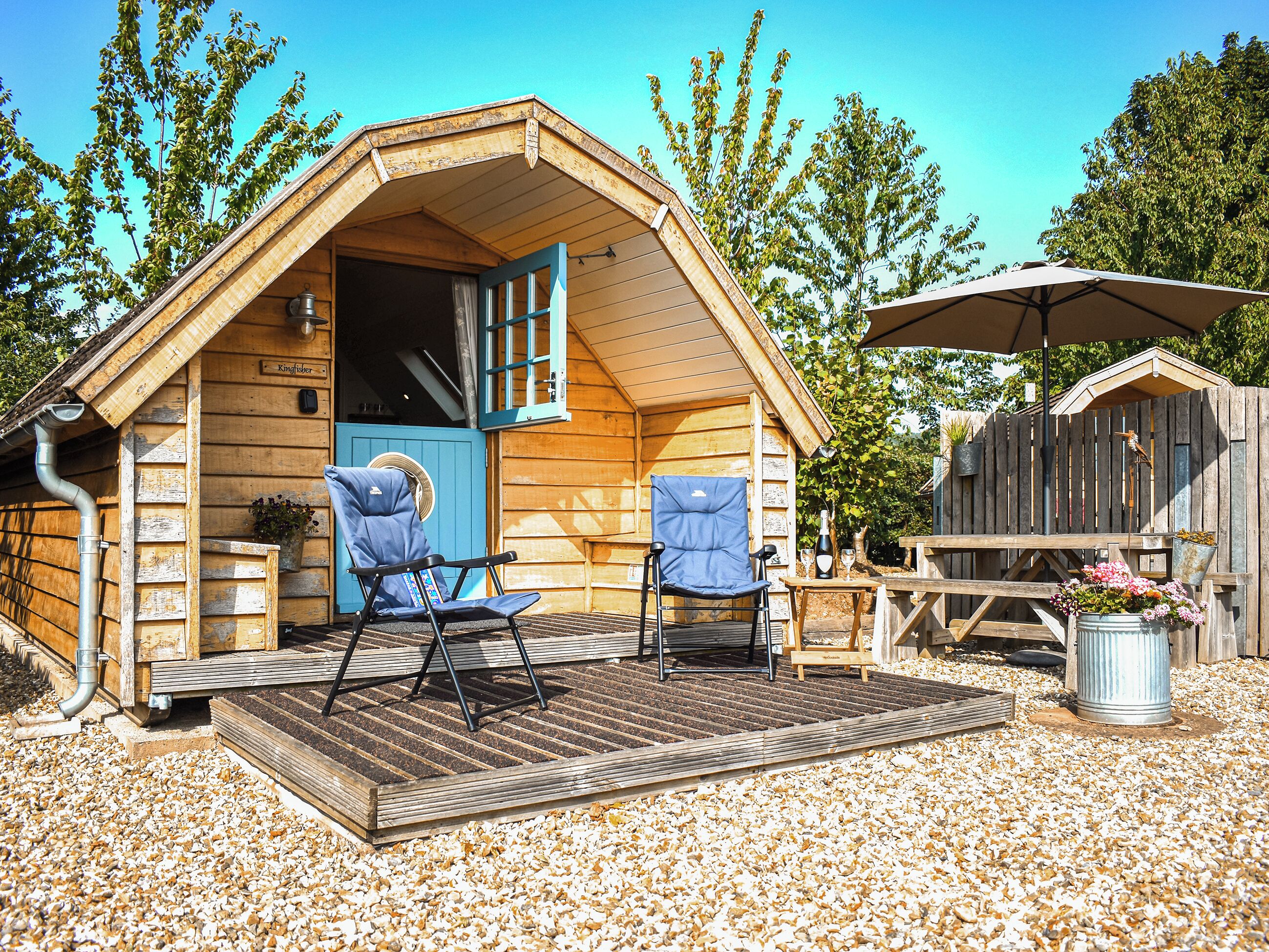 A wooden cabin with a pitched roof, a blue door, and two blue chairs on a gravel patio.
