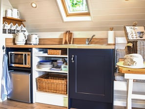 A compact kitchen with a microwave, shelves with dishes, a sink, and a small table with a hat. - 1 bedroom accommodation in Sidmouth (Sidmouth)