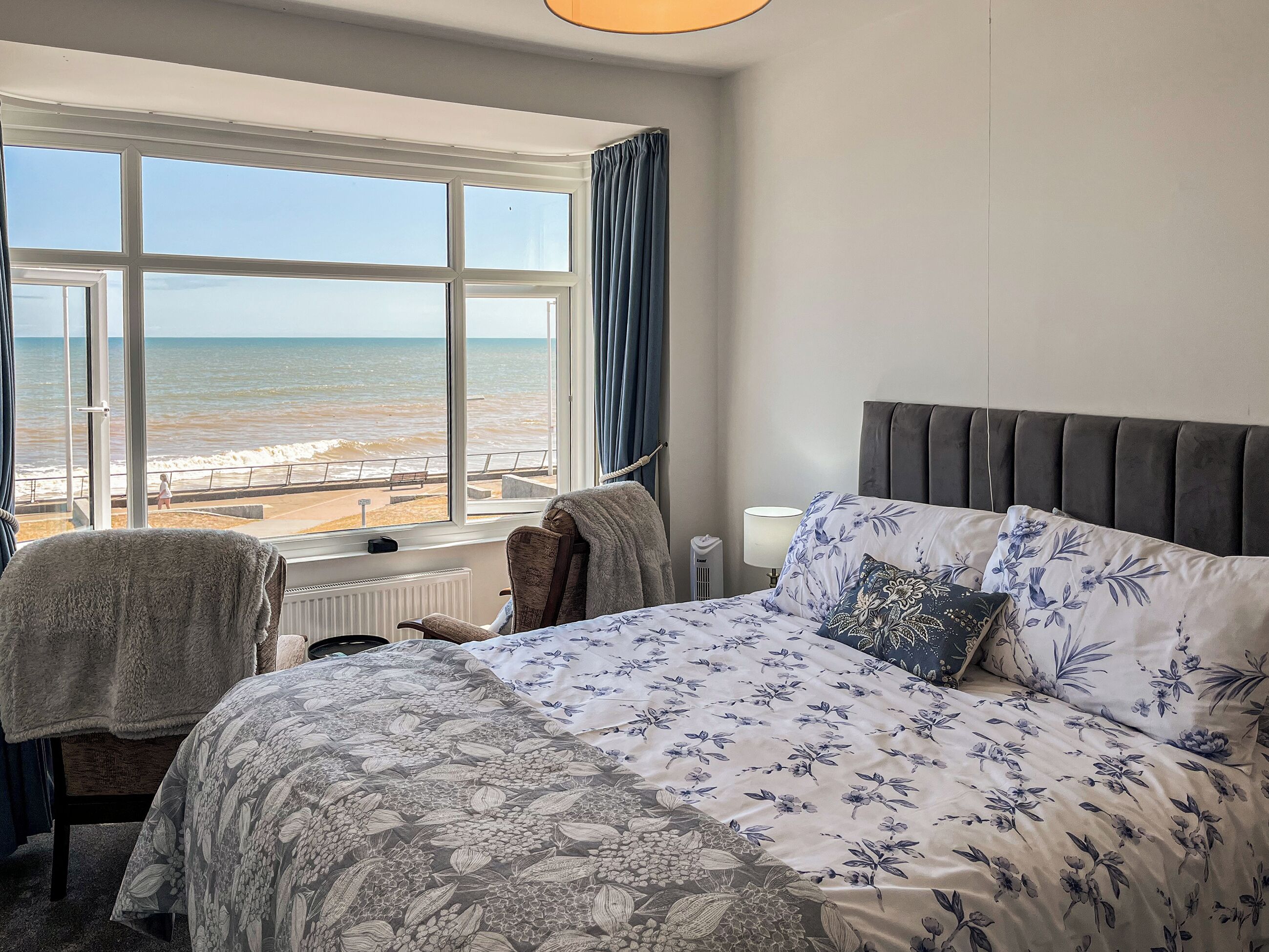 Ein Schlafzimmer mit großem Fenster mit Blick auf den Strand, einem Bett mit floraler Bettwäsche, einem Stuhl mit Handtuch und einem Nachttisch mit Lampe.