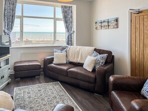 A living room with a brown leather sofa, a white radiator, a patterned rug, and a view of the sea through a window with patterned curtains. - The sea views from this property are amazing and the beach is right out front. (Hornsea)