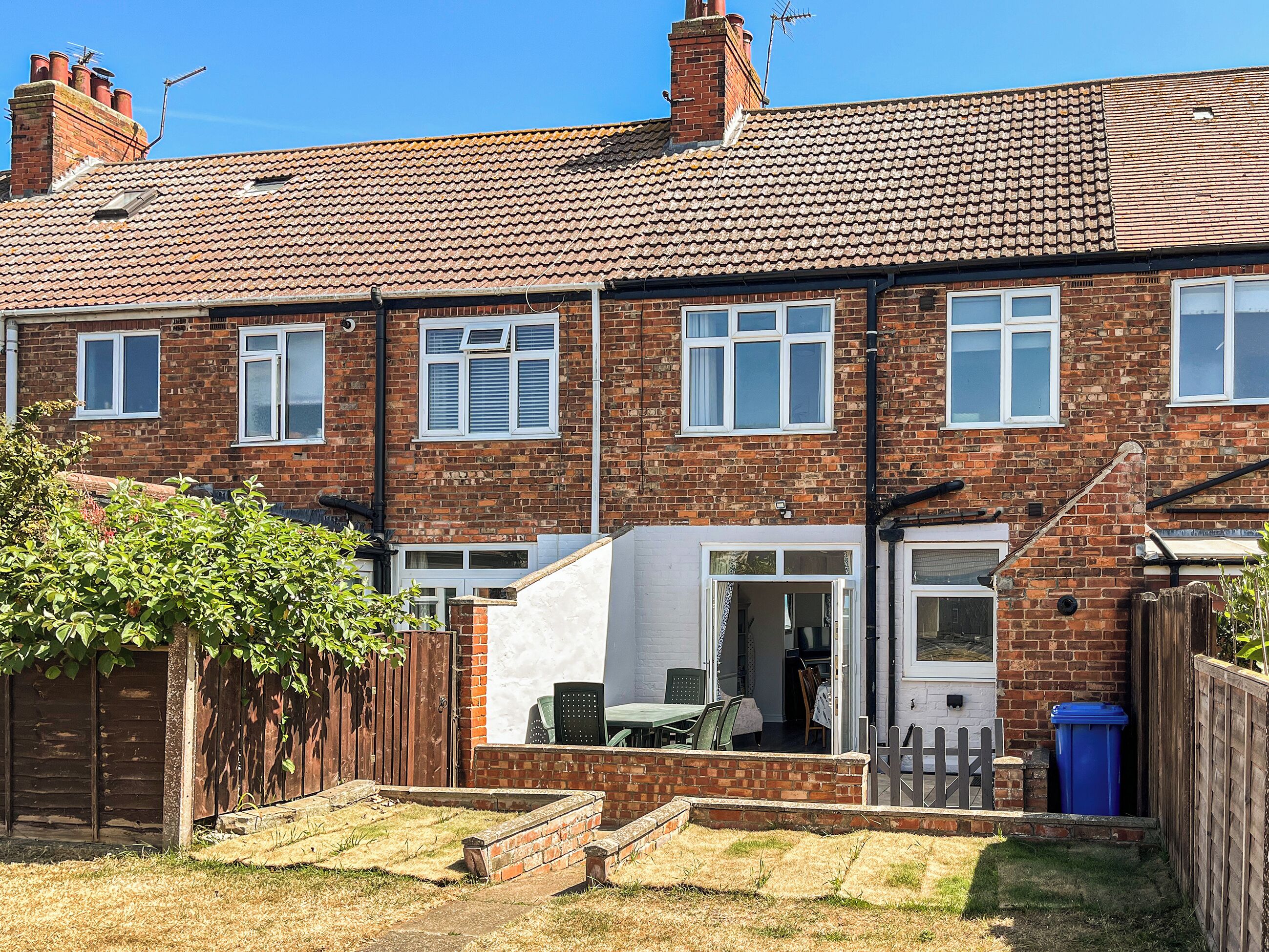 A two-story brick terraced house with a front garden, a visible door, and a window.