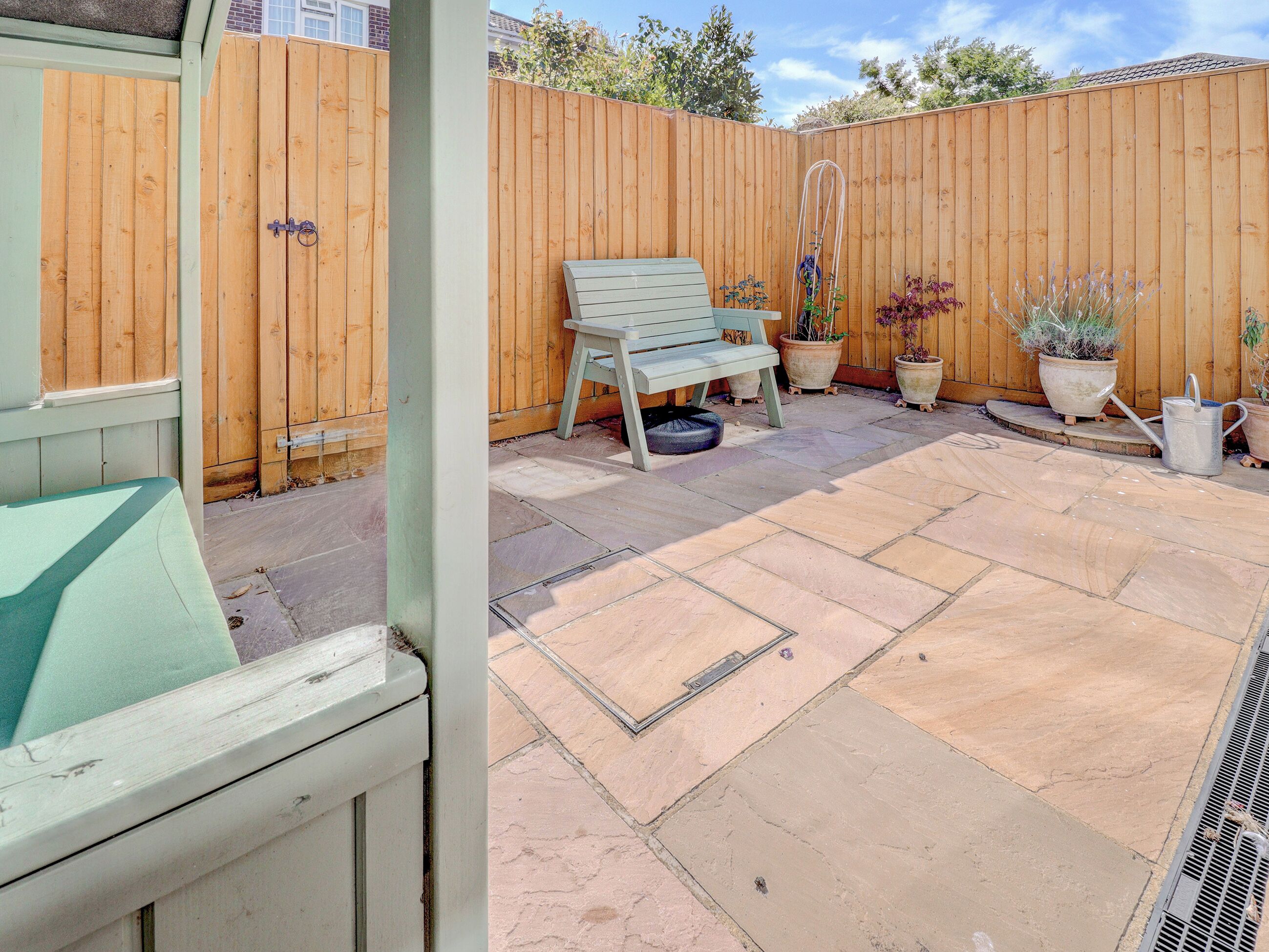 A patio with a wooden fence, a bench, potted plants, and a metal watering can.