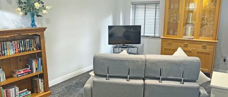 Ein Wohnzimmer mit grauem Sofa, einem Holzregal, einem Flachbildfernseher und einer Holzkabine mit Glastüren.