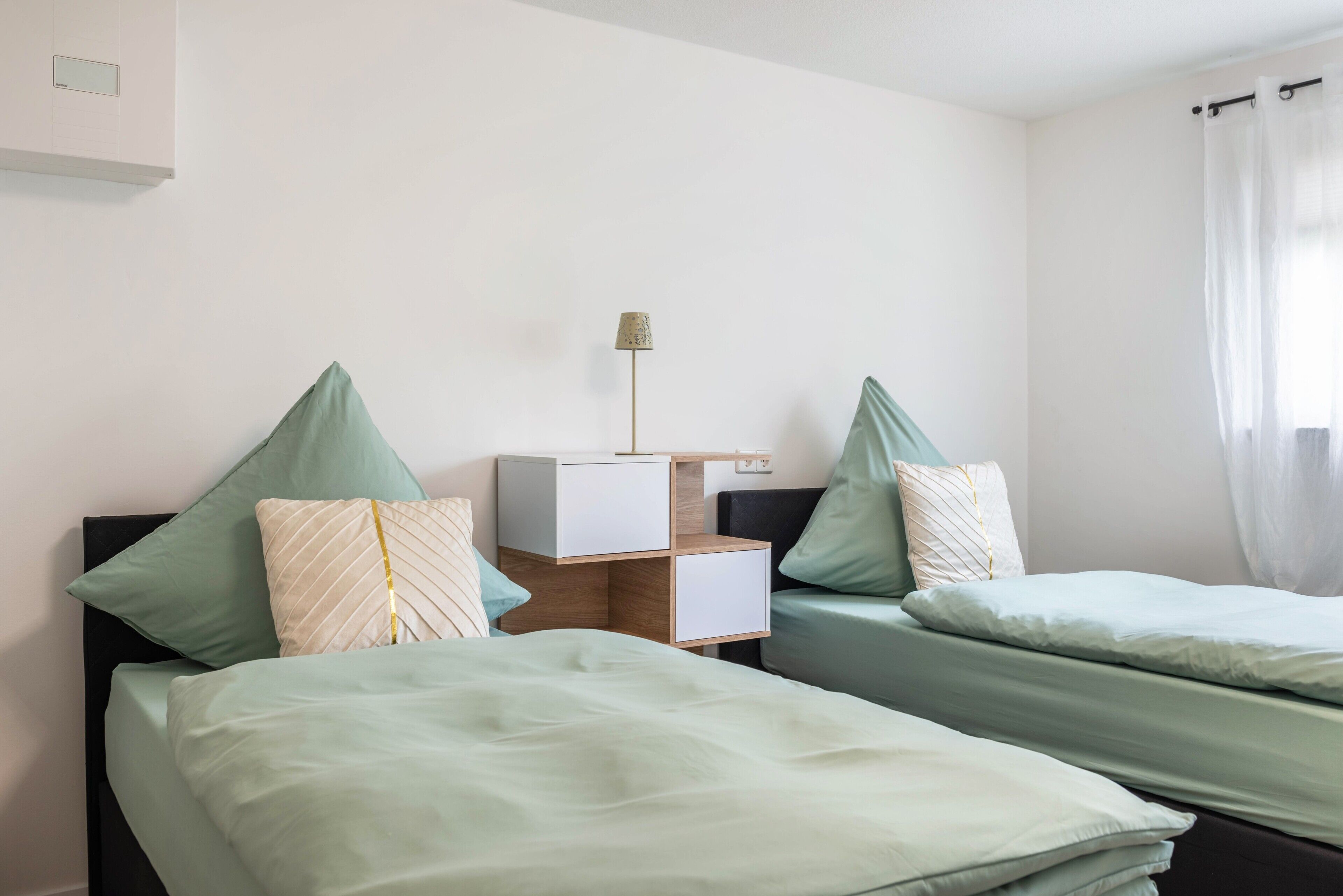 Quarto de hotel com duas camas, uma mesa de cabeceira, uma luminária e uma janela com cortinas.