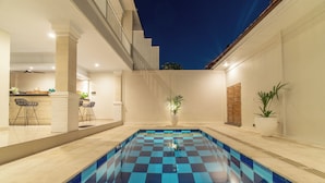 Una piscina con diseño de baldosas de ajedrez, rodeada por un patio con mobiliario y plantas en macetas.