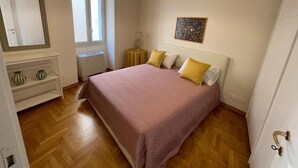 A bedroom with a bed, a window, a bedside table, a lamp, and a painting on the wall. - Foro Romano Apartments E (Rome)