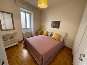 A bedroom with a bed, a window, a bedside table, a lamp, and a painting on the wall.