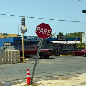 Vista da rua com uma placa de pare vermelha, fachada azul de um estabelecimento e uma caminhonete vermelha.