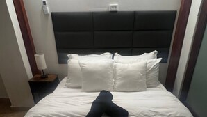Een hotelkamer met een bed, kussens, een nachtkastje met een lamp en een bedieningspaneel aan de muur.