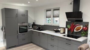 A modern kitchen with grey cabinets, a built-in oven, a coffee machine, and a window with blinds. - Modern Apartment “Frankenruhe” near Bamberg on the Edge of Franconian Switzerland (Strullendorf)